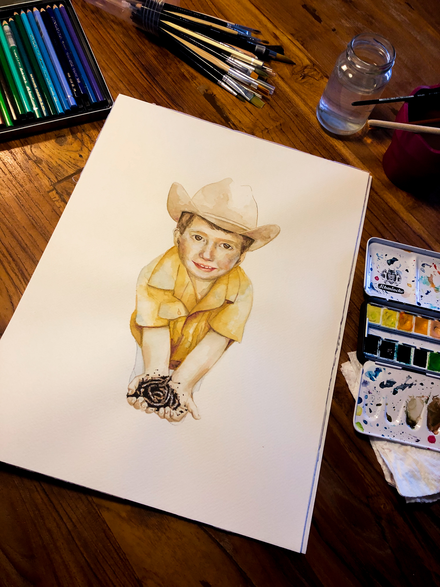 Zu sehen ist ein Aquarell-Porträt eines kleinenJungen mit Cowboyhut und gelben Hemd. Erhält Regenwürmer in den Händen und guckt den Betrachter direkt an.